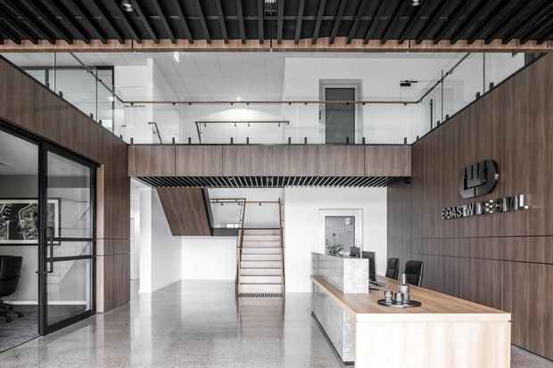 Contemporary commercial reception interior with a double-height void and mezzanine level. The space features dark timber-look wall panelling, a black exposed slatted ceiling, glass balustrades and partitions, polished concrete or terrazzo-like flooring, and a light timber reception desk with a stone-faced counter. A stair leads to the upper level, while the company logo is mounted on the feature wall.