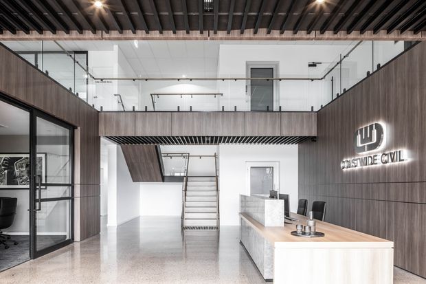 Modern commercial reception and office interior with a double-height atrium, mezzanine level, exposed dark ceiling battens, glass balustrades, dark timber wall panelling, polished concrete flooring, and a light timber reception desk. A staircase and glazed meeting or office rooms are visible.