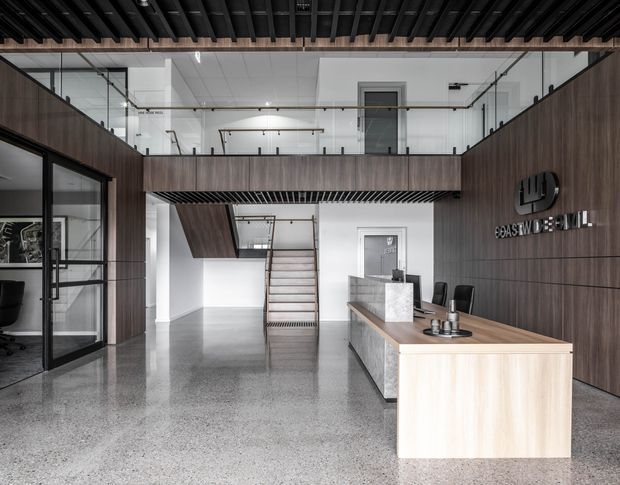 A contemporary commercial reception and lobby interior with a double-height atrium, polished concrete or terrazzo-style flooring, dark timber wall panelling, glazed office partitions, an upper-level mezzanine with glass balustrades, and a timber reception desk with stone counter. A stair rises to the mezzanine, while black exposed ceiling structure and linear detailing create an industrial-modern character.