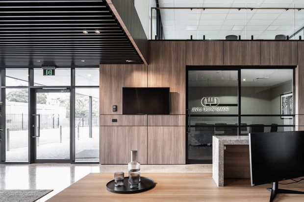 A contemporary commercial reception or waiting area with a timber-look feature wall, wall-mounted television, black-framed glass partitions, a reception counter, and a polished light floor. The ceiling features exposed black linear battens and suspended services, while large glazed entry doors provide bright natural light.