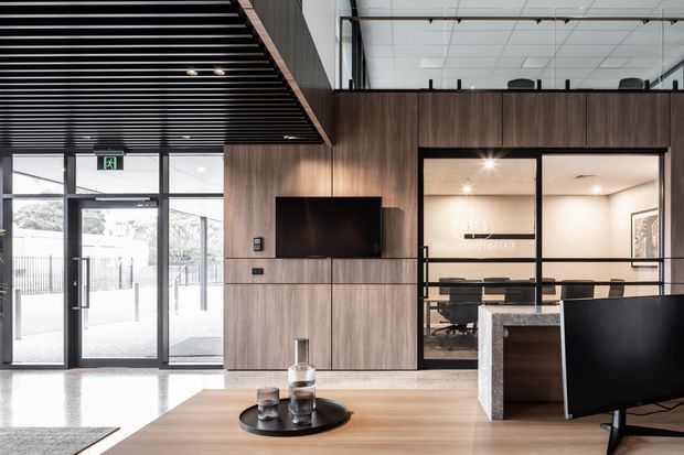 Contemporary commercial interior reception or lounge area with dark timber wall panelling, integrated television, polished concrete flooring, a timber coffee table, and a glass-enclosed meeting room. A black linear slatted ceiling and exposed upper glazing add an industrial-modern character, while the entrance glazing brings in daylight.