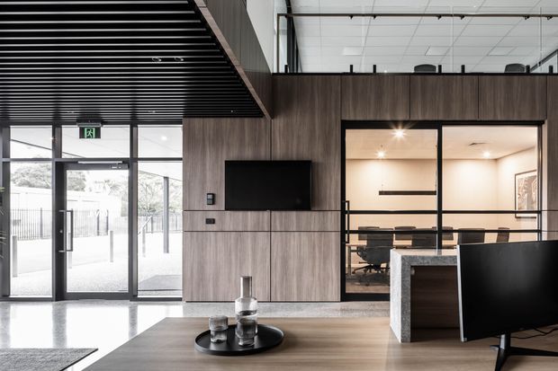 A contemporary commercial office reception or lounge area with dark timber-look wall panelling, integrated television, polished concrete flooring, and a low timber coffee table. Black-framed glazed doors and partitions open to a meeting room with a conference table and chairs. The space features a black linear slatted ceiling, exposed services, a reception counter, and a bright industrial-modern fit-out.
