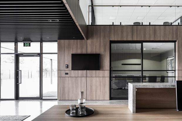 A contemporary commercial reception or lobby with dark timber wall panelling, integrated television, black-framed glass meeting room, stone reception counter, polished concrete flooring and a black slatted ceiling. A mezzanine or upper-level workspace is visible above the glazed partition. The space has a refined, minimalist corporate fit-out with strong charcoal, timber and stone tones.