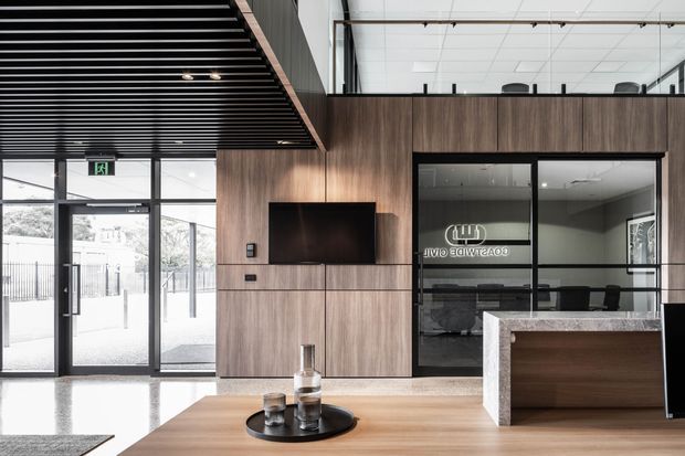 Contemporary commercial reception or lobby with a timber-lined feature wall, integrated television, reception desk, glass-fronted meeting room, polished concrete floor, and black slatted ceiling. Abundant glazing and an open mezzanine-like upper level create a bright, professional interior.