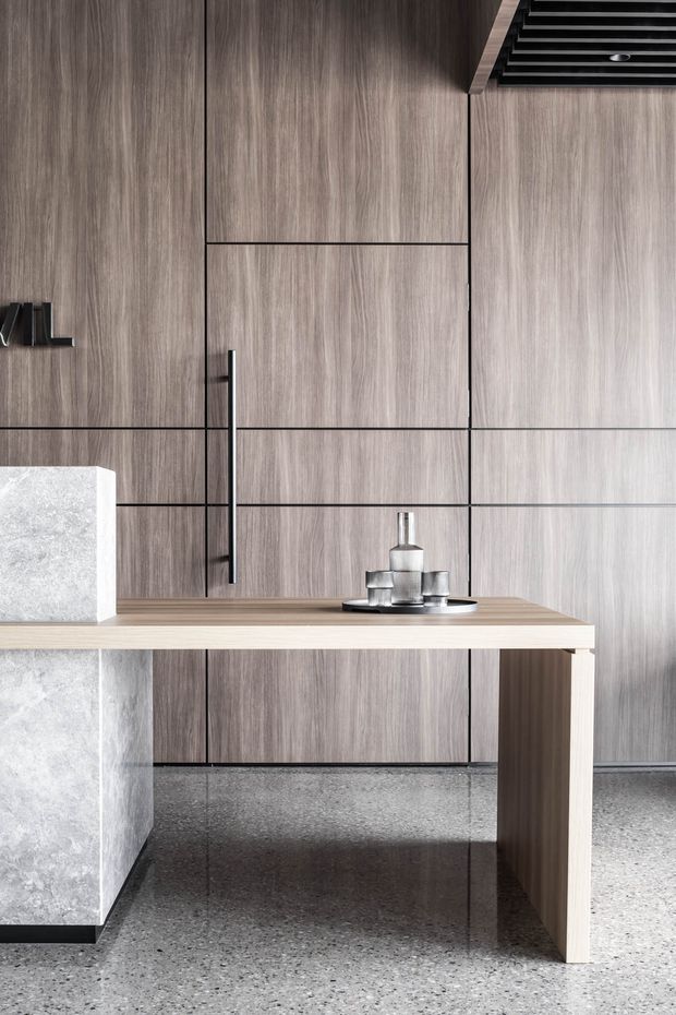 Close-up view of a refined contemporary kitchen featuring full-height vertically grained wood-look cabinetry with dark linear reveals and a long slim black handle. A pale stone island or counter with a light timber overhang and supporting leg occupies the foreground, paired with a polished grey terrazzo floor. Minimal metallic objects sit on the counter, and a black ceiling ventilation grille is visible above.