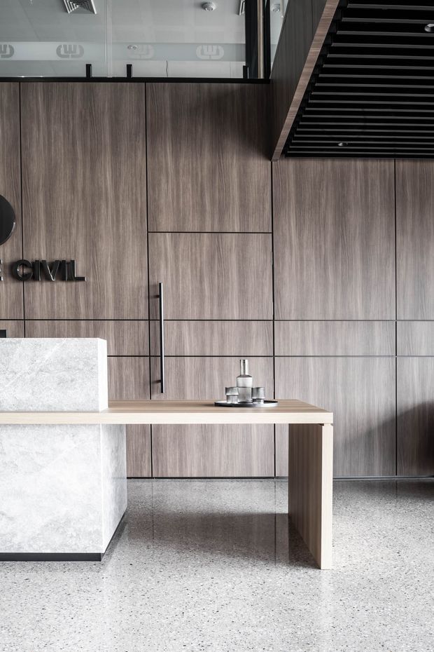 A refined contemporary commercial reception or workspace interior featuring full-height grey-brown timber-look wall panelling and integrated cabinetry, a pale stone-clad reception counter with timber return, and a terrazzo-style floor. Black linear ceiling detailing, slim black framing, and minimal signage add a crisp architectural character.