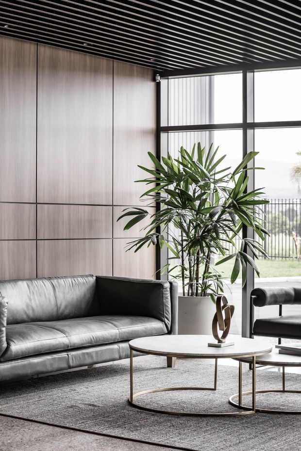 A refined contemporary living room with a large grey leather sofa, round white coffee table, dark metal bases, and a matching lounge chair. The room features full-height timber wall panelling, a black linear slatted ceiling, expansive black-framed glazing, a large indoor palm, and a neutral textured rug. The overall aesthetic is sophisticated, monochromatic, and softly lit.