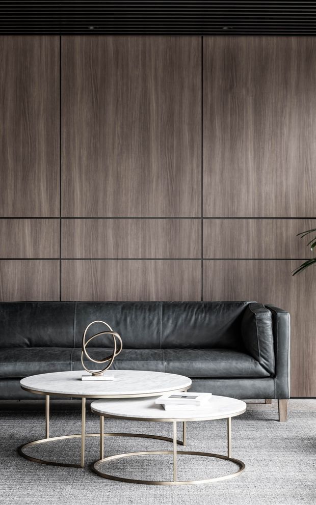 A refined contemporary living room vignette featuring a black leather sofa against a full-height dark timber wall with fine black grid joints. Two overlapping round coffee tables with pale stone tops and slender metallic bases sit on a light textured rug, complemented by a sculptural circular ornament and minimal books. A dark slatted ceiling and a small leafy plant add depth to the moody, sophisticated interior.