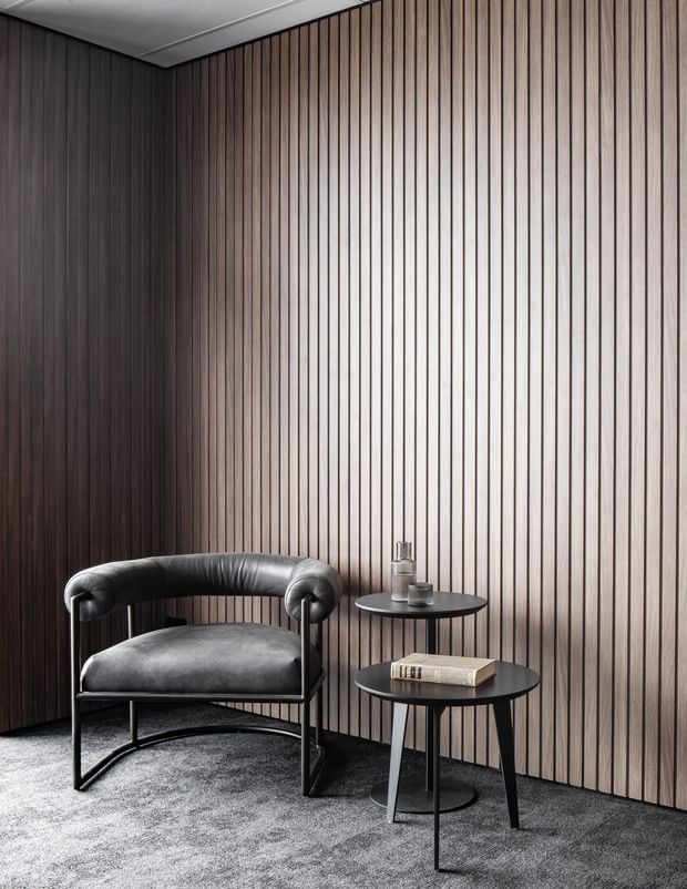 A refined, minimalist interior corner featuring a black leather lounge chair and two round black side tables against a full-height vertical timber slat wall. The room has dark grey carpet flooring, subdued lighting, and a clean white ceiling, creating a sophisticated, textural composition.