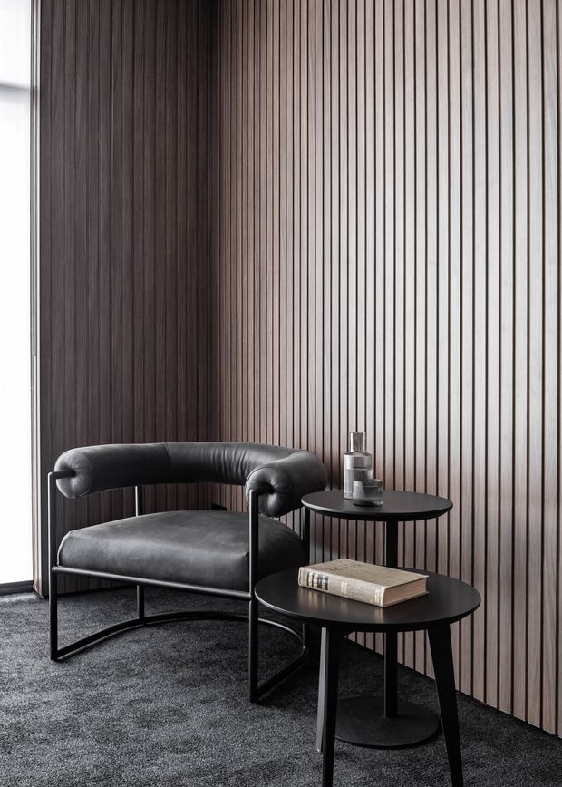 A refined seating vignette with a black leather lounge chair, two round black side tables, and a book, set against a full-height dark timber vertical slat feature wall. A grey carpet and narrow window edge complete the quiet, monochromatic interior.