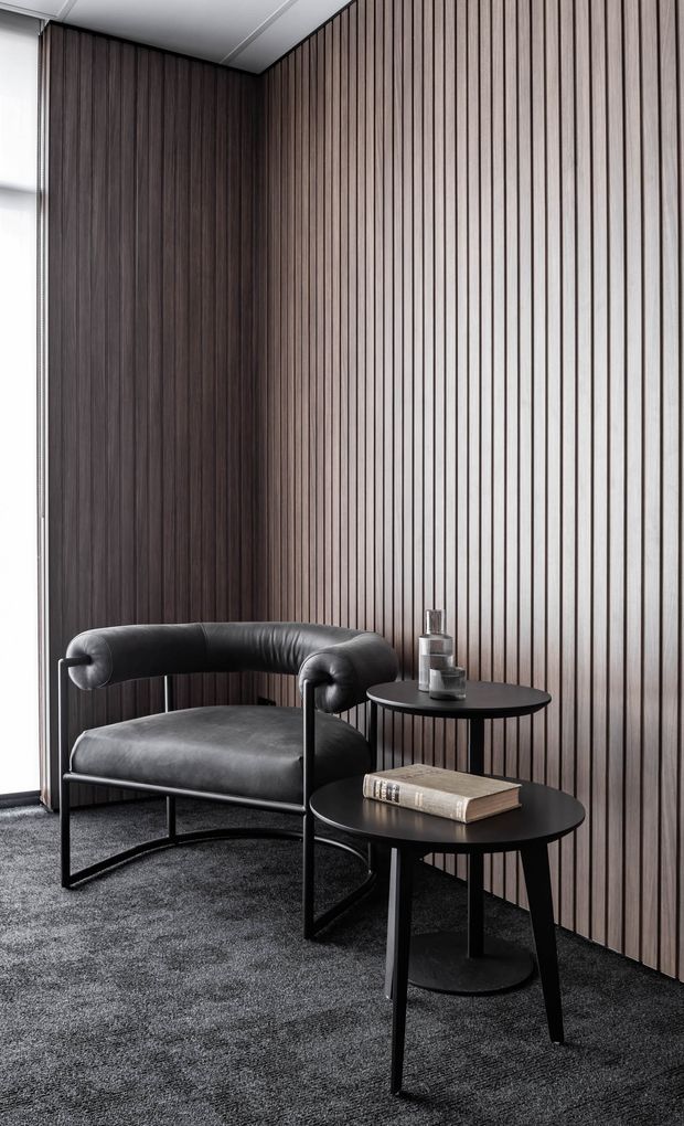 A refined seating nook featuring a black leather lounge chair, two round black side tables, and dark textured carpet. The space is enclosed by a dramatic vertical timber slat feature wall in dark brown tones, with a narrow window at the left and a simple white ceiling. The styling is minimalist, sophisticated, and monochromatic with strong vertical rhythm.