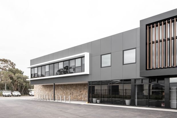 Contemporary commercial building exterior with a dark grey panelled facade, extensive black-framed glazing, a recessed vertical timber screen, and a contrasting stone-clad lower level. The building fronts a paved forecourt with trees and parked vehicles in the background.