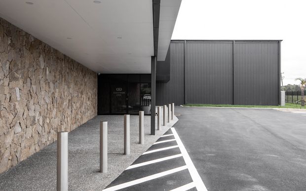 Exterior architectural view of a modern commercial or institutional building entry and covered drop-off area. The facade combines rough natural stone cladding with dark charcoal vertical metal cladding, a flat projecting canopy, recessed downlights, and a row of pale bollards beside a striped pedestrian safety zone and asphalt forecourt.