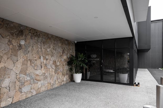 Modern commercial entrance beneath a covered canopy, featuring a rough natural-stone feature wall, dark metal-framed glazed entry doors, charcoal vertical metal cladding, and a light aggregate concrete forecourt. Potted plants flank the entrance.