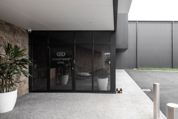 Exterior entrance to a contemporary commercial building, featuring a covered entry canopy, dark aluminium-framed glazed doors and windows, rough stone wall cladding, and a large corrugated metal boundary wall. Potted plants flank the glass entrance, with a paved forecourt and stainless-steel bollards.