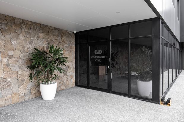 Entrance to a contemporary commercial building with a rough natural-stone feature wall, black-framed glazed doors and windows, a white soffit, and a textured grey forecourt. Potted plants flank the entry, and the glazing displays Coastwide Civil branding.