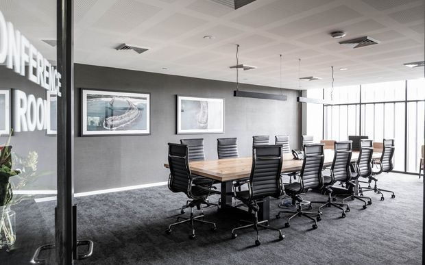A contemporary commercial conference room with a long timber meeting table surrounded by black ergonomic office chairs. The room has dark grey walls, charcoal carpet, framed artwork, extensive floor-to-ceiling glazing, suspended linear lighting, and a white acoustic-tile ceiling. A glass entry partition with partial lettering is visible on the left.