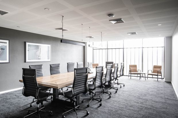 Bright contemporary commercial meeting room with a long light-wood boardroom table, black ribbed office chairs, a linear suspended pendant light, grey carpet, dark feature wall, framed artwork, and extensive floor-to-ceiling glazing. The room has a suspended acoustic ceiling with recessed lighting and overlooks an exterior area through the windows.