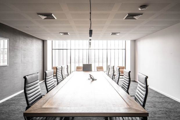A modern commercial conference room with a long timber meeting table surrounded by black and white office chairs. The room has grey carpet, a textured grey feature wall, white walls, a suspended ceiling with recessed lighting and HVAC vents, and extensive floor-to-ceiling glazing at the far end.