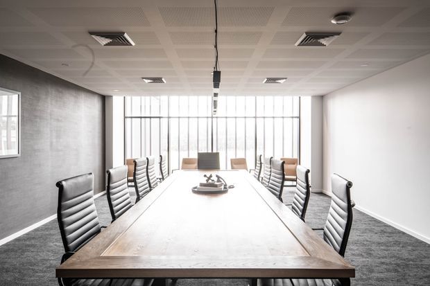 A bright contemporary conference room with a long timber meeting table, multiple black leather chairs, grey carpet, a textured grey feature wall, and full-height industrial-style glazing at the far end. The ceiling has acoustic panels, recessed services, and suspended lighting.
