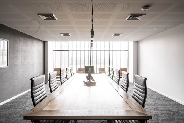 Bright contemporary boardroom with a long timber conference table, rows of black-and-white office chairs, grey carpet, a textured dark grey feature wall, and expansive floor-to-ceiling glazed windows. The suspended linear light and acoustic tiled ceiling reinforce the formal commercial workspace setting.
