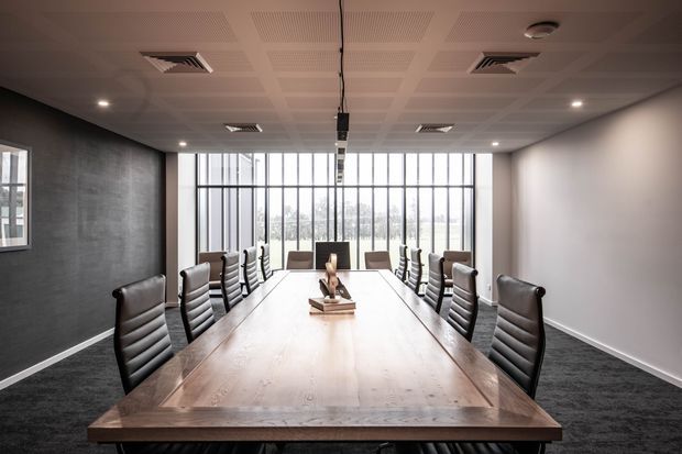 A contemporary commercial boardroom with a long solid-wood conference table surrounded by black padded office chairs. The room has dark carpet flooring, grey textured feature walls, white walls, a suspended acoustic ceiling with recessed lighting and linear air-conditioning vents, and full-height glazed windows at the far end.