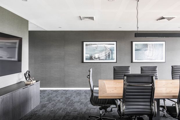 A refined modern dining and meeting room with a long timber table surrounded by black upholstered office chairs. The room features dark textured wall finishes, a charcoal carpet, a wall-mounted television above a dark built-in credenza, framed artwork, and a white acoustic ceiling with recessed services and suspended linear lighting.