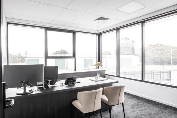 Bright contemporary office or reception workspace with a long dark built-in desk, computer monitors, telephone, and two upholstered visitor chairs. The room has expansive floor-to-ceiling corner glazing with dark frames, a suspended white acoustic tile ceiling, and dark carpet flooring.