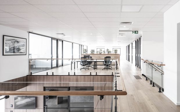 Bright contemporary commercial office interior viewed from an upper-level landing, with an open-plan workstation area, glass partitions and balustrades, light timber flooring, timber-clad stair or void surround, exposed grid ceiling, and abundant glazing. The space has a clean, airy, professional fit-out with a visible stair opening and corridor circulation.