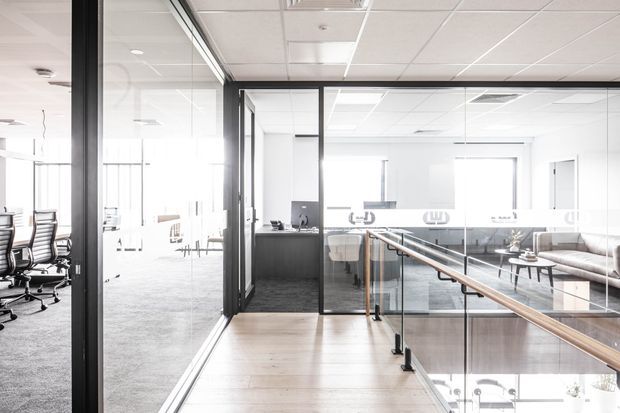 Bright contemporary commercial office interior with glass-partitioned meeting rooms, open workspaces, a reception or workstation area, and a glazed balustrade overlooking a lower level. The space features white acoustic ceiling tiles, dark metal framing, pale timber flooring, grey carpet, and minimalist office furniture.