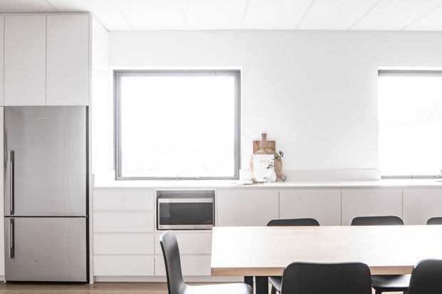 Bright, minimalist kitchen and dining area with pale flat-panel cabinetry, a stainless-steel refrigerator, integrated microwave, long light benchtop, and a dining table with dark upholstered chairs. Large windows provide abundant natural light, while simple white surfaces and a flush tiled or panelled ceiling create a clean contemporary interior.