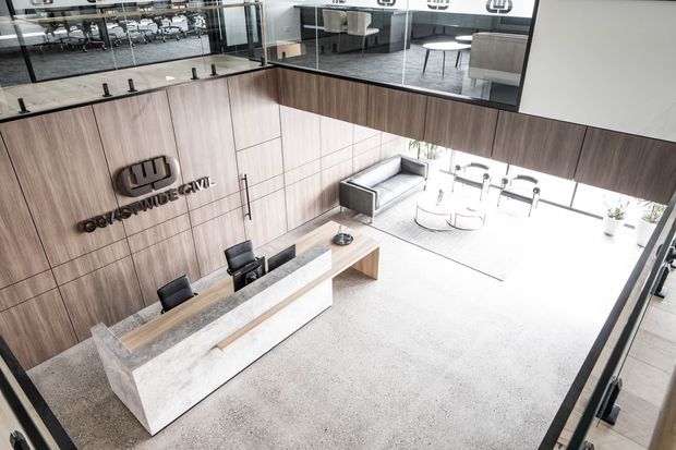 A bright, contemporary commercial reception and lounge area viewed from an elevated angle. A long pale stone reception desk with dark office chairs sits against a warm timber-panelled feature wall with raised lettering. Behind it are glazed upper-level meeting or office spaces, while the lower lounge includes a grey sofa, round coffee tables, and abundant natural light. The space has a refined neutral palette and polished professional fit-out.
