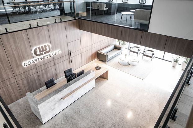 A bright, double-height commercial reception lobby viewed from above. The space features a long stone reception desk, warm vertical timber wall panelling with illuminated company signage, polished terrazzo flooring, black-framed glass meeting rooms or offices above and behind, and a small lounge seating area with abundant glazing.