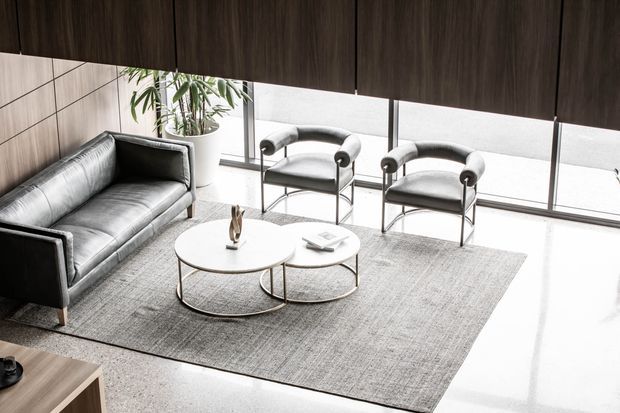 A refined contemporary living room viewed from above, featuring a charcoal leather sofa, two matching sculptural lounge chairs, nested round coffee tables, and a large textured grey rug. Floor-to-ceiling glazing provides abundant natural light, while dark timber wall panelling and a warm timber cabinet frame the space.