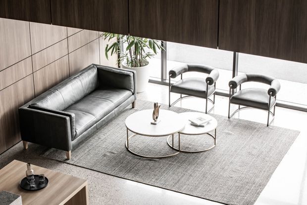 A sophisticated contemporary living room with a grey leather sofa, two sculptural lounge chairs, nested round coffee tables, and a large textured area rug. Dark timber wall panelling and overhead cabinetry contrast with bright floor-to-ceiling glazing, creating a refined, minimalist seating area.