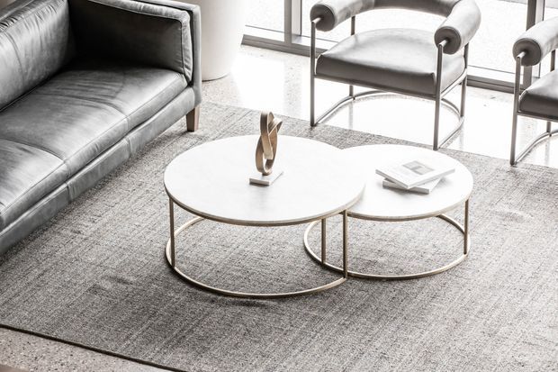 Bright contemporary living room vignette featuring overlapping round nesting coffee tables with slim metallic frames and pale tops, a grey leather sofa, matching upholstered lounge chairs, and a large textured grey rug beside expansive glazing. The palette is refined and neutral with strong natural light.