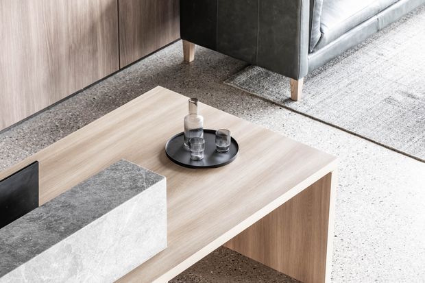Close-up of a refined contemporary living room featuring a pale timber coffee table with an integrated grey stone block, a black circular tray with glassware, grey upholstered seating, grey textured carpet, and a dark textured stone or concrete partition element.