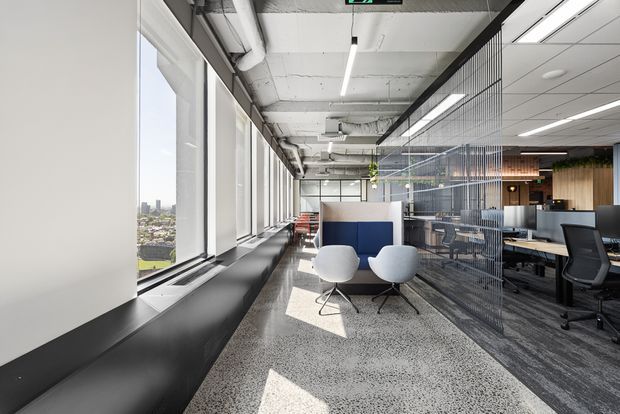 A bright contemporary commercial office interior with expansive floor-to-ceiling windows, exposed concrete services and ceiling structure, polished terrazzo flooring, and an open-plan workstation area screened by a black metal mesh partition. A small lounge setting with two white chairs sits beside a blue upholstered divider, while timber-look joinery and greenery add warmth in the background. The urban skyline is visible through the windows.