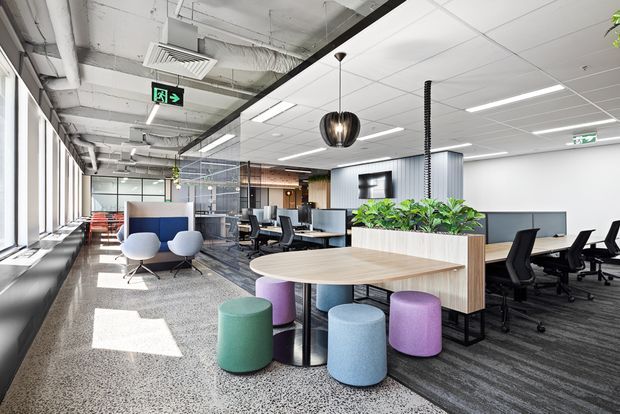 A bright contemporary open-plan commercial office with exposed services, extensive glazing, shared workstations, acoustic partitions, and a communal high table with colourful upholstered stools. Indoor planting, polished terrazzo flooring, carpeted work areas, and pendant lighting create a lively collaborative workspace.