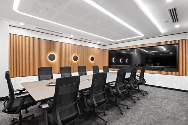 Contemporary boardroom with a long rectangular conference table surrounded by black ergonomic office chairs. The room features a warm vertical timber-slat feature wall, integrated circular wall lights, a large wall-mounted display, white marble-look lower wall panels, grey carpet, and a recessed linear LED ceiling design.