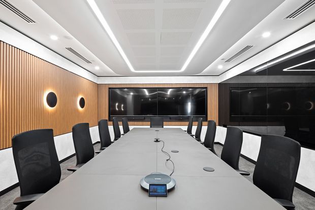 A sophisticated modern conference room with a long light-coloured meeting table, black mesh-backed chairs, integrated video-conferencing equipment, large dark display screens, warm vertical timber wall panelling, and sculptural linear LED ceiling lighting. The room has a high-contrast black, white, and warm timber palette.