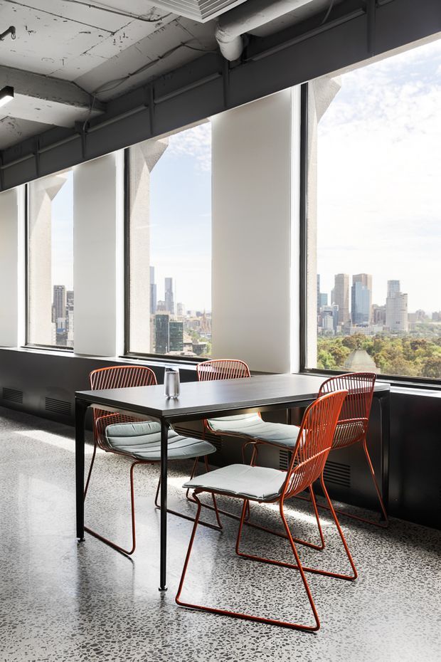 A bright urban dining area in an industrial-style interior, with a black rectangular table surrounded by four orange-red wire-frame chairs with light cushions. Large floor-to-ceiling windows provide expansive city views, while exposed services, dark ceiling structure, black window frames, and polished terrazzo flooring reinforce the contemporary commercial aesthetic.