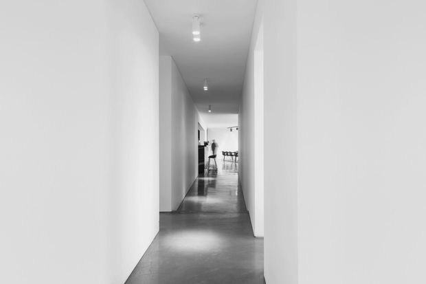 A long, narrow minimalist hallway with crisp white walls, recessed ceiling lights, and a smooth dark grey polished floor. The corridor opens toward a brighter dining or living area in the distance, creating a strong linear perspective and uncluttered monochrome appearance.