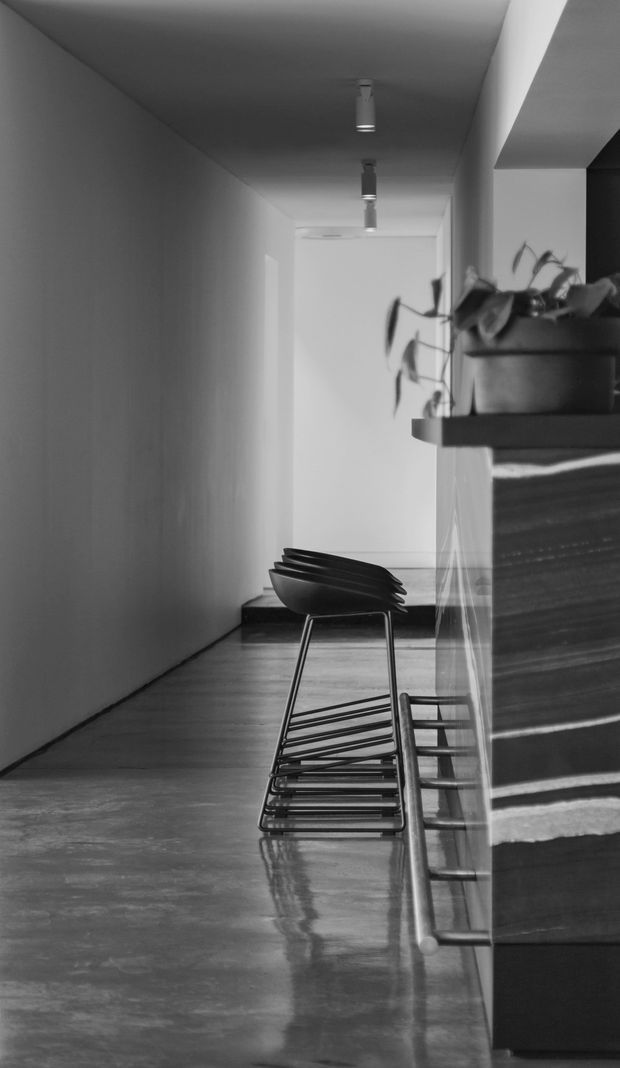 A monochrome photograph of a narrow, minimalist interior passage beside a kitchen or bar counter. The space features polished concrete flooring, smooth white walls and ceiling, recessed and surface-mounted linear lighting, dark timber or stone cabinetry, a projecting shelf with a potted plant, and a row of black sculptural stools. Reflections on the glossy floor and cabinetry add depth to the composition.