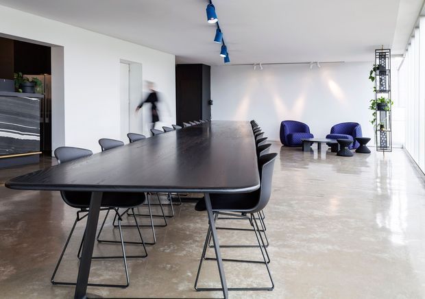 A spacious contemporary dining and meeting area with a long dark dining table surrounded by black chairs. The room has polished concrete flooring, white walls, dark built-in cabinetry, track lighting, vivid blue pendant lights, and a small blue lounge seating arrangement beside floor-to-ceiling glazing. A narrow black metal plant shelving unit stands by the windows.