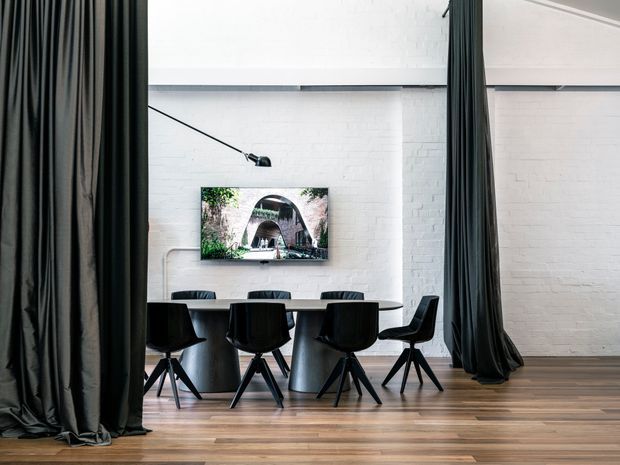A refined dining area framed by full-height dark curtains, featuring a long dark dining table with black chairs, white-painted brick walls, exposed dark ceiling beams, timber flooring, and a wall-mounted artwork or screen. A slim black wall light extends over the table, creating a dramatic, gallery-like interior.