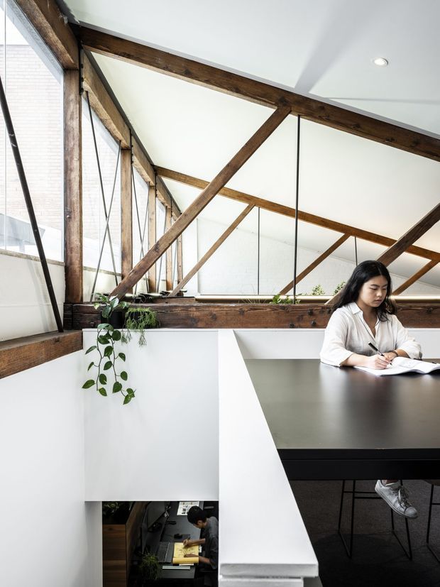 A bright contemporary loft-style workspace with a large dark table, a person writing, and exposed rustic timber trusses and diagonal bracing beneath a sloped ceiling. White walls, expansive glazing, black-framed window elements, indoor plants, and a view down to a lower-level work area create an airy mezzanine office with a strong connection to exposed structure.