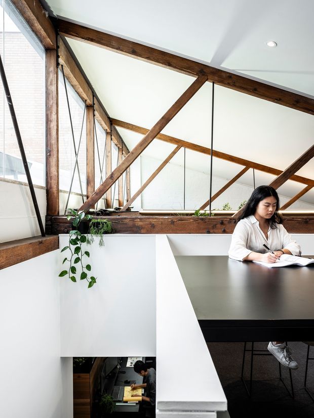 A bright contemporary loft-style workspace with a large dark tabletop, white walls, exposed reclaimed timber roof framing, and extensive internal glazing. A person is seated at the desk, while a lower-level workspace is visible through the open void. Potted trailing plants add greenery to the otherwise restrained interior.