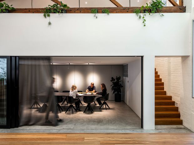 Interior view of a contemporary multi-level home featuring a sunken or enclosed dining area behind large black-framed glazed sliding doors. The dining space has a dark table, chairs, pendant-style track lighting, and concrete flooring. A timber staircase rises beside a white-painted brick wall, while the foreground has warm timber flooring and the upper level includes exposed timber beams with trailing plants.