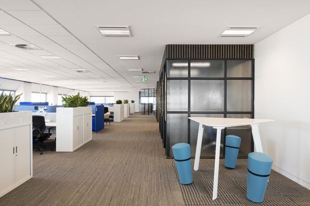 Bright open-plan commercial office interior with rows of workstations, white storage cabinets, blue office chairs, carpeted flooring, and abundant glazing. A black-framed translucent glass partition encloses a casual meeting or breakout zone with a white high table and blue stools. Suspended acoustic ceiling tiles and recessed fluorescent lighting create a clean, functional workplace environment.